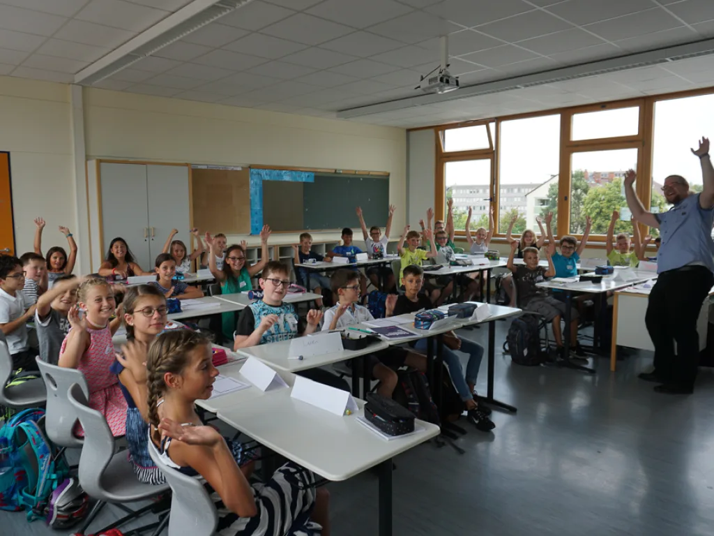Kinder melden sich, die Lehrkraft zeigt nach vorn.